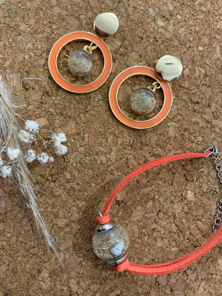 Plan sur un ensemble de bijoux. Sur un fond en liège, une paire de boucle d'oreille façonnant un anneau rond orange avec une belle en verre remplie de sable à l'intérieur de l'anneau, suivis d'un bracelet en en cordon de cuir brossé orange, accessoirisé d'une bulle en verre remplir de sable avec un coquillage. Les éléments sont en mouvement à l'intérieur des bulles.